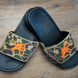 A pair of Old Skool Camo Camp Slides with a waterproof PVC camo upper and orange detailing on a wooden floor.