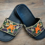 A pair of Old Skool Camo Camp Slides with a waterproof PVC camo upper and orange detailing on a wooden floor.