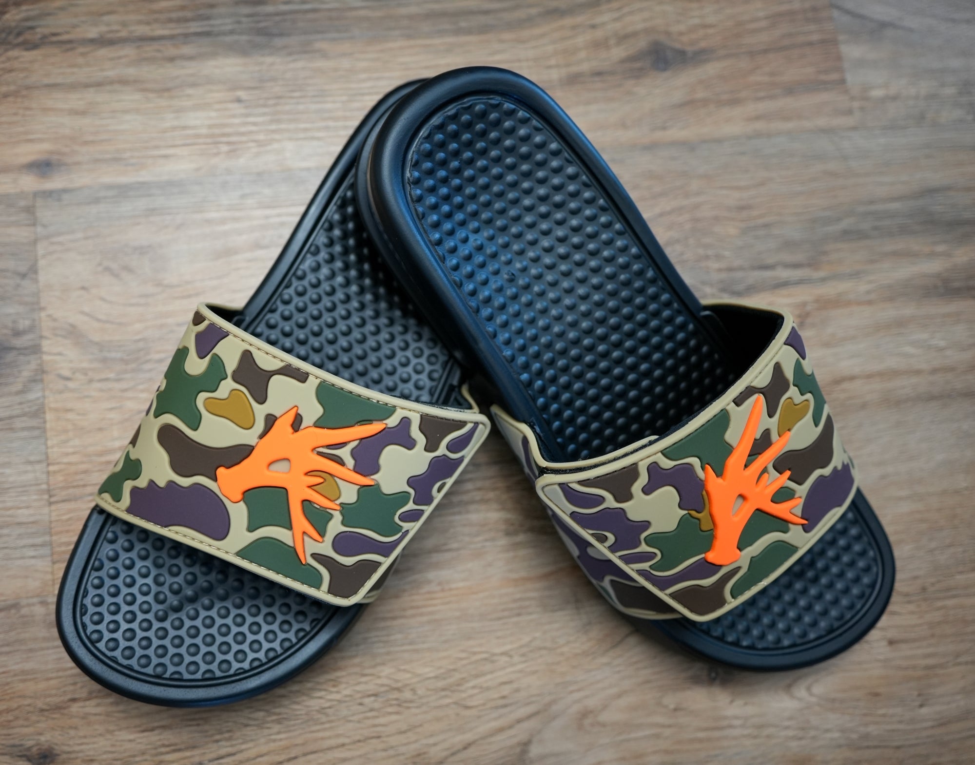 A pair of Old Skool Camo Camp Slides with a waterproof PVC camo upper and orange detailing on a wooden floor.